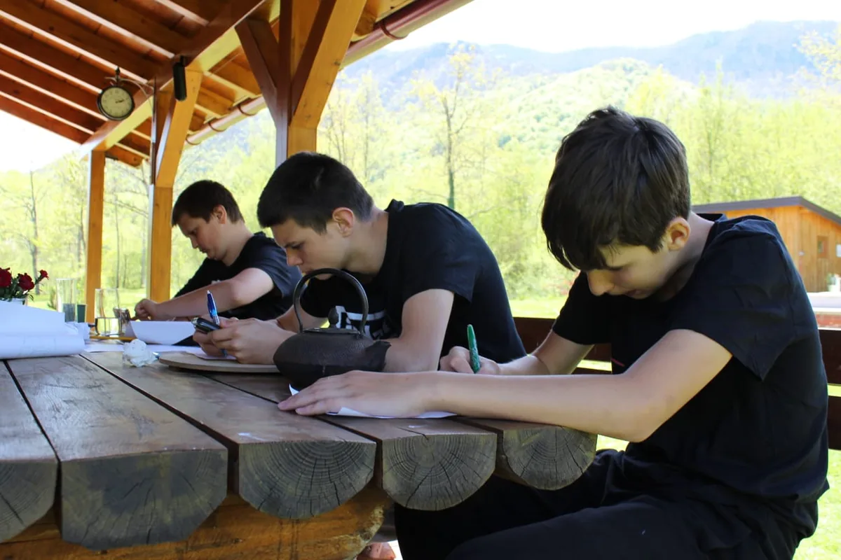 Participants writing at a wooden table under a shelter, with mountains behind