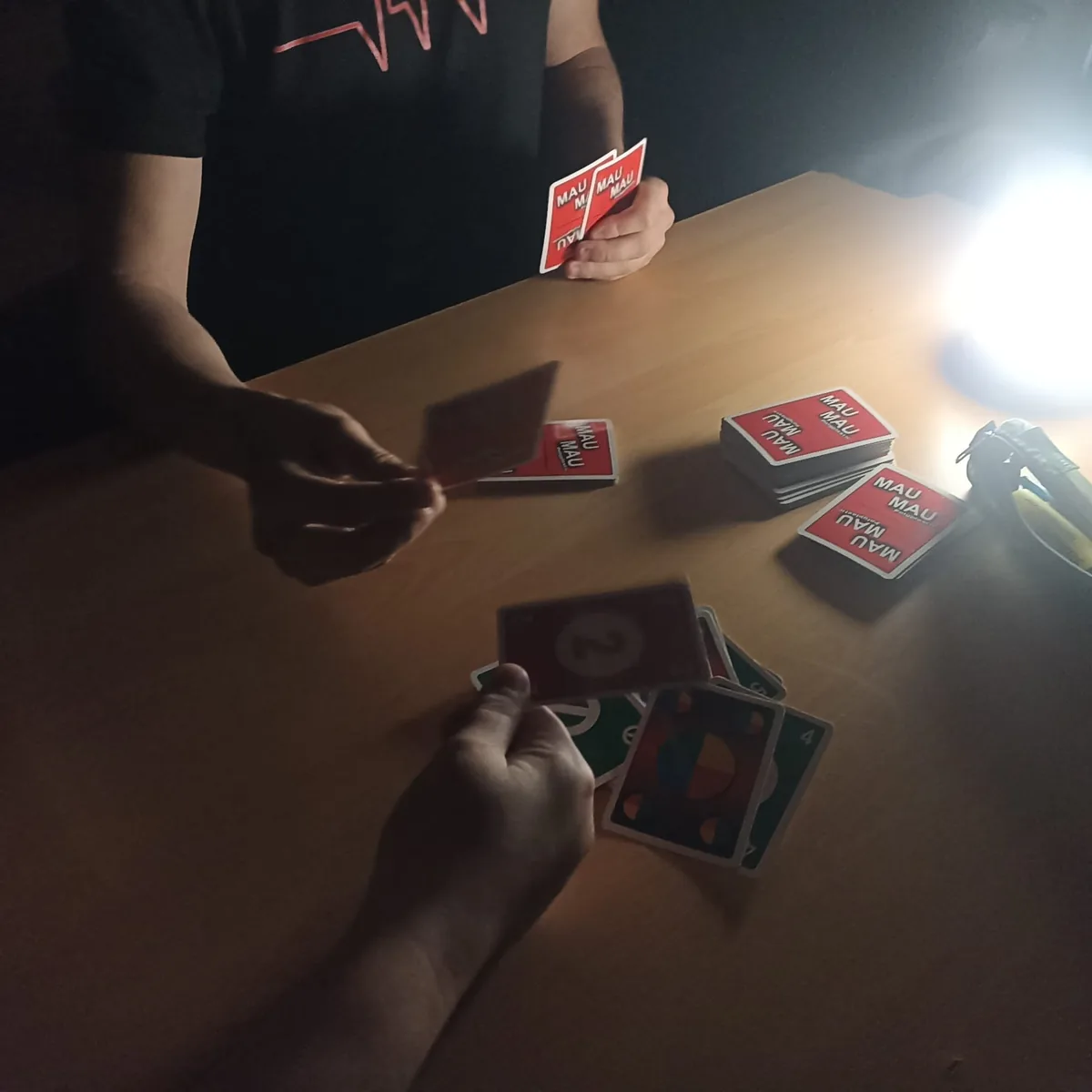 Hands holding MAU MAU playing cards at a table under a lamp