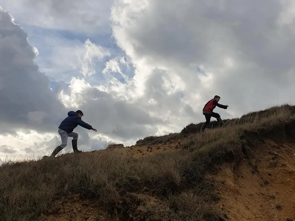 Two participants climbing a grassy hill under clouds